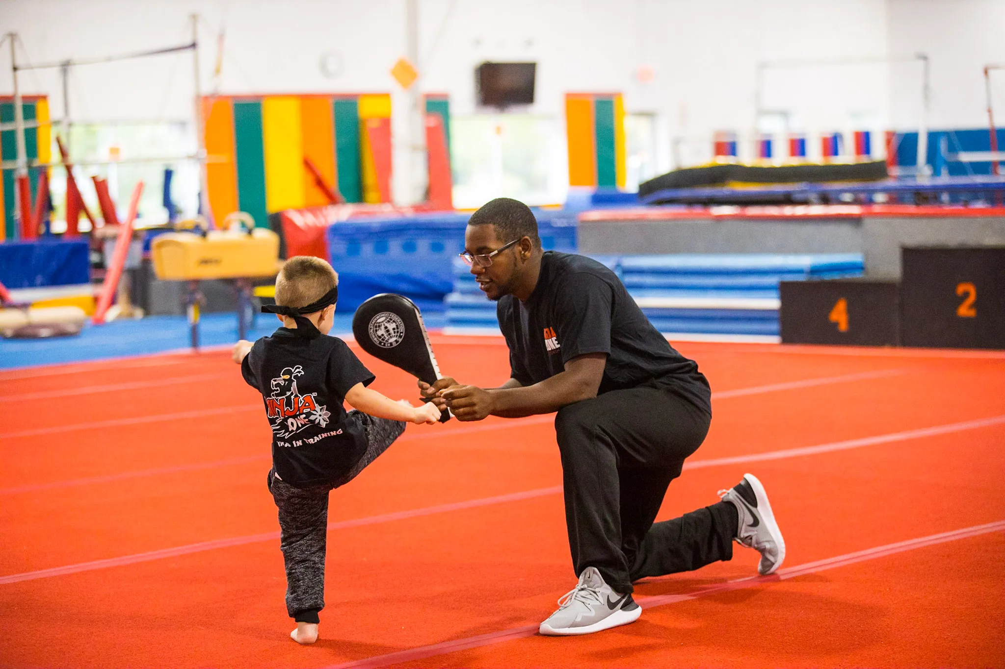 An EGA coach kneels to spot a young ninja student on the orange tumble track